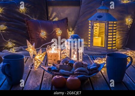 Coin salon confortable avec café et biscuits sur un balcon, décoré pour l'hiver avec des lanternes, bougies, couvertures en laine et des lumières de fées dans le beau soleil Banque D'Images
