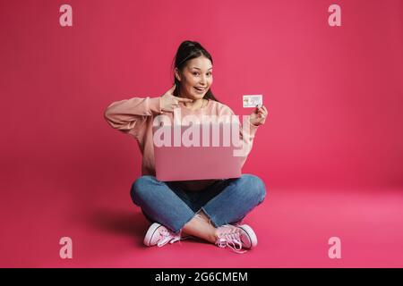 Femme souriante asiatique pointant du doigt sur la carte de crédit tout en utilisant un ordinateur portable isolé sur le mur violet Banque D'Images