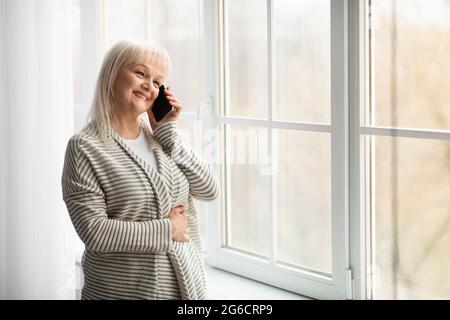 Femme mûre souriante parlant au téléphone à la maison près de la fenêtre Banque D'Images