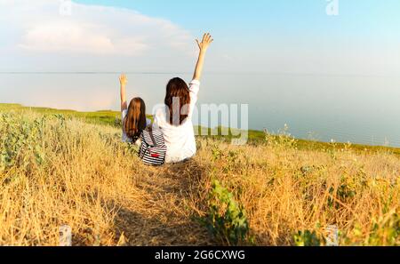 Vue arrière d'une femme méconnaissable qui embrasse une fille tout en étant assise herbe séchée et paysage pittoresque pendant le week-end d'été Banque D'Images