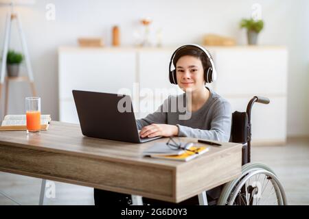 Enseignement à domicile. Adolescent gai handicapé en fauteuil roulant portant des écouteurs, ayant une leçon en ligne sur ordinateur portable à l'intérieur Banque D'Images