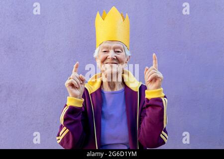 Content athlète féminine senior dans les vêtements de sport et couronne de papier pointant vers le haut avec les doigts tout en regardant l'appareil photo sur fond violet Banque D'Images