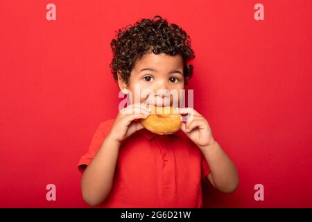 Adorable enfant aux cheveux bouclés mangeant un beignet savoureux et regardant l'appareil photo sur fond rouge Banque D'Images