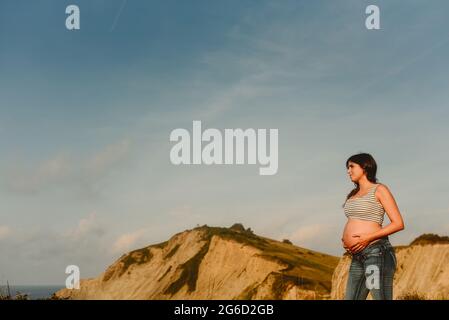 Vue latérale de la jeune femme enceinte hispanique paisible en jeans et crop top révélant le ventre debout sur le bord de mer contre les montagnes Banque D'Images