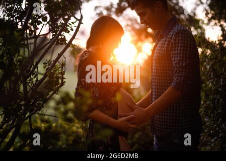 Vue latérale d'un jeune couple heureux s'attendant à un bébé debout face à face et touchant le ventre contre la lumière du coucher de soleil dans la forêt verte Banque D'Images