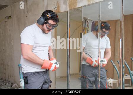 Le constructeur change un foret sur le chantier de construction. Travailleur de la construction portant des gants de sécurité, des lunettes et des couvre-oreilles. Banque D'Images