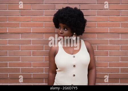 Les jeunes afro ont un modèle afro-américain en robe et boucles d'oreilles pendu contre le mur de briques rouges et regardant loin Banque D'Images