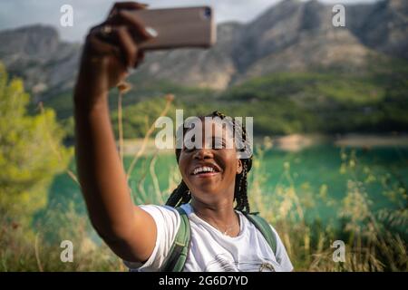 Une femme afro-américaine ravie de prendre sa photo sur son smartphone sur fond de lac dans les montagnes en été Banque D'Images