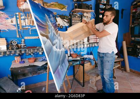 Homme barbu dans des vêtements décontractés enlever le papier protecteur de la toile avec paysage abstrait pendant le travail en studio créatif Banque D'Images