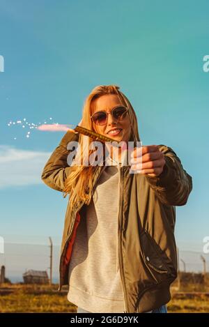 Petit angle de la femme blonde souriante dans des lunettes de soleil tenant une bougie étincelante dans la campagne avec le ciel bleu Banque D'Images