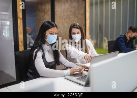 Des collègues multiraciaux portent un masque assis à la table avec un ordinateur portable et discutent de projets professionnels tout en travaillant dans un espace de travail Banque D'Images