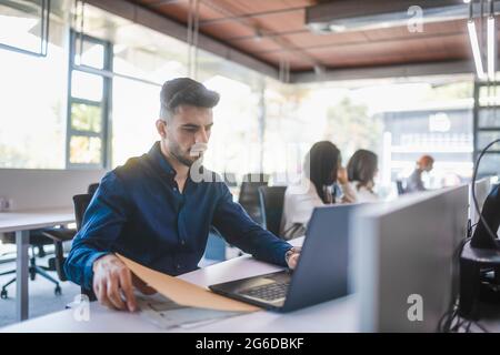 Employé masculin concentré assis à une table avec un ordinateur portable et lisant des documents tout en travaillant dans un bureau spacieux avec des collègues flous Banque D'Images