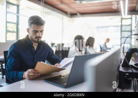 Employé masculin concentré assis à une table avec un ordinateur portable et lisant des documents tout en travaillant dans un bureau spacieux avec des collègues flous Banque D'Images