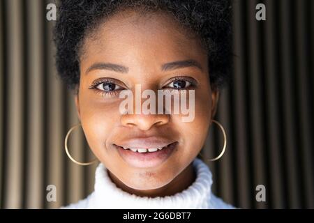 Joyeux élégant jeune femme noire en pull court regardant l'appareil photo tout en se tenant près du mur de bâtiment rayé dans la ville Banque D'Images