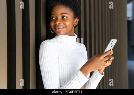 Bonne jeune femme noire élégante en surfant sur un smartphone tout en se tenant près du mur de bâtiment rayé de la ville Banque D'Images