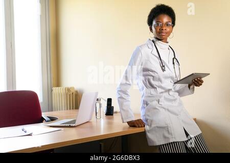 Positive jeune femme noire médecin dans un manteau médical et des lunettes avec stéthoscope regardant la caméra tout en travaillant avec une tablette dans un bureau de clinique moderne Banque D'Images