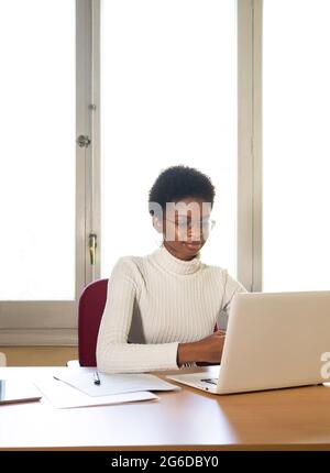 Femme afro-américaine dactylographiant rapport sur ordinateur portable tout en travaillant à table dans le bureau de clinique moderne Banque D'Images