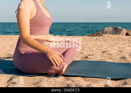 Vue latérale d'une petite femme sereine non reconnaissable assise à Padmasana et faisant du yoga tout en méditant avec les yeux fermés sur la mer de sable Banque D'Images