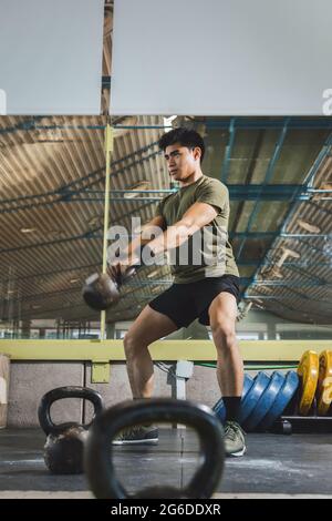 Homme asiatique s'entraîner sur les épaules et les bras avec de lourdes kettlebells dans la salle de gym pendant l'entraînement fonctionnel et regarder loin Banque D'Images