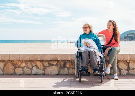 Fille adulte gaie avec une mère âgée en fauteuil roulant, une clôture en pierre le long de la promenade près de la mer en été Banque D'Images