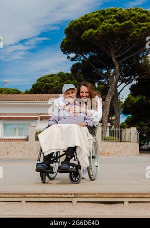 Femme adulte souriante et homme âgé en fauteuil roulant se détendant ensemble en ville par beau temps Banque D'Images