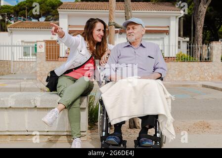 Femme adulte souriante et homme âgé en fauteuil roulant se détendant ensemble en ville par beau temps Banque D'Images