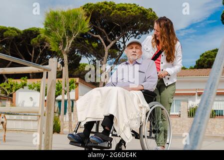 Femme adulte souriante et homme âgé en fauteuil roulant se détendant ensemble en ville par beau temps Banque D'Images