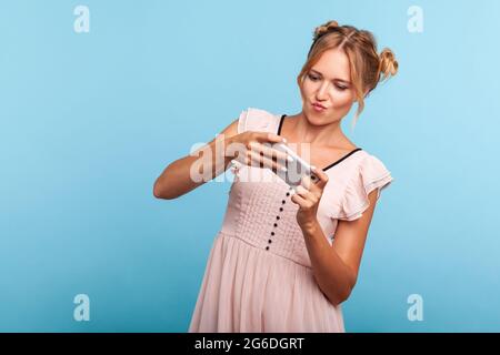 Portrait d'adorable femme blonde avec deux faisceaux de cheveux utilisant un téléphone portable avec une expression ciblée attentive, jouant à un jeu vidéo sur un téléphone portable. Intérieur Banque D'Images