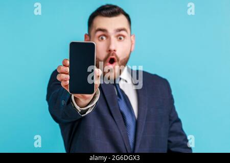 Portrait d'un homme émerveillé en costume élégant tenant le téléphone portable et regardant l'appareil photo avec les yeux grands et la bouche ouverte, en disant wow surpris au sujet de la technologie Banque D'Images