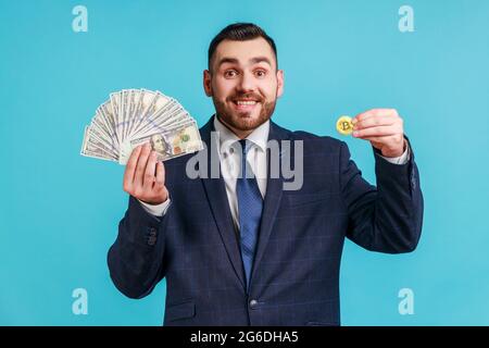 la crypto-monnaie et l'argent. Portrait d'un homme à la barbe riche et heureux costume élégant montrant des billets de banque en dollars et un bitcoin doré, souriant à l'appareil photo. Intérieur s Banque D'Images