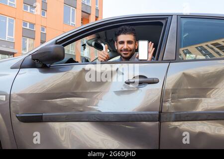 Portrait d'un homme barbu dans une voiture écrasée regardant l'appareil photo et souriant, montrant le pouce vers le haut et le smartphone avec écran vide, publicité inspection d'assurance Banque D'Images