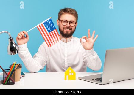 Heureux homme souriant tenant le drapeau des États-unis d'amérique et montrant ok geste avec les doigts, assis sur le lieu de travail, satisfait de se déplacer à l'étranger. Indoo Banque D'Images