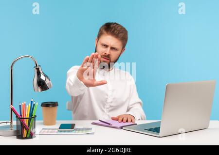 Homme d'affaires strict avec barbe assis dans le bureau avec ordinateur portable montrant l'arrêt, avertissement avec le geste de la main, accès refusé, interdiction. Indoo Banque D'Images