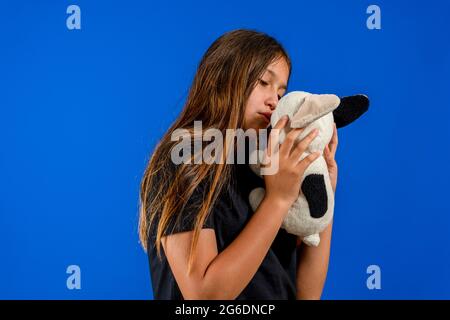 Photo de studio de petite fille avec son jouet de chien doux dans les mains, debout contre le mur bleu portant une chemise rayée, écharpe et chapeau avec pompon, regardant le guide Banque D'Images