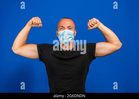 Jeune homme beau en masque médical et t-shirt jaune isolé sur fond bleu. Personne forte montrant le muscle de bras, confiant et fier de pouvoir à d Banque D'Images