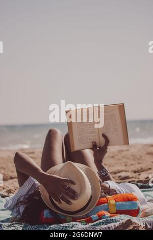 Vue arrière verticale femme indienne noire couchée sur la plage lisant un livre alors qu'elle tient son chapeau d'été lors d'une journée d'été ensoleillée et venteuse. Vibes d'été 2021 Banque D'Images