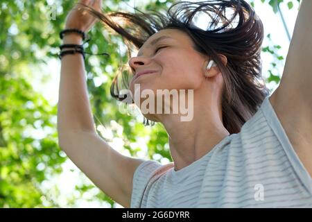 Une femme heureuse écoute de la musique dans des écouteurs sans fil et danse, avec ses cheveux en vol, un jour ensoleillé d'été.