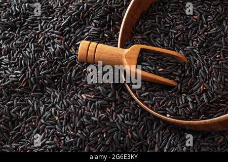 Une pelle en bois brune dans un bol en bois rempli de grains de riz noirs secs crus sur fond de graines de riz noires. Riz noir biologique pour une alimentation diététique. Banque D'Images
