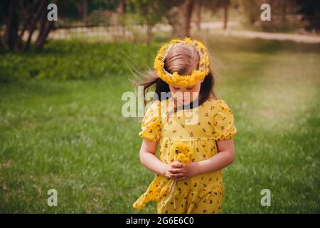 une jolie fille aux cheveux amples et une couronne de pissenlits jaunes sur sa tête regarde un bouquet de pissenlits avec sa tête abaissée dans un beau vert Banque D'Images