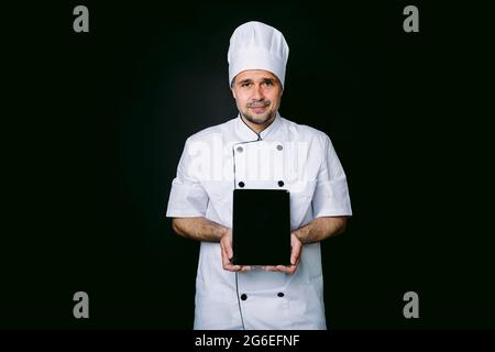 Chef cuisinier portant une veste et un chapeau de cuisine, tenant une tablette, montrant son écran, sur fond noir Banque D'Images