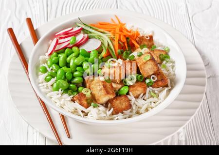 bol à poke de tofu avec riz basmati, haricots édamames, radis, concombre finement coupé et carottes dans un bol blanc avec baguettes, vue horizontale de l'abov Banque D'Images