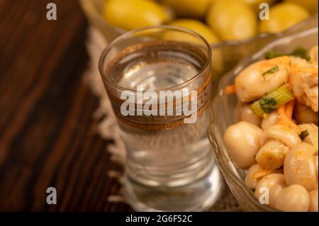 Un verre de vodka et de champignons salés et des olives salées dans des plats en verre sur fond de tissu de maison avec une texture rugueuse. Gros plan sélectif Banque D'Images