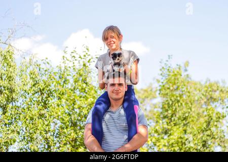 Frère défoqué de la sœur de circonscription à l'arrière. Portrait d'une fille heureuse sur les épaules de l'homme, porcgyback. Fille tenant chat, chaton. Famille jouant à l'extérieur. PET, an Banque D'Images