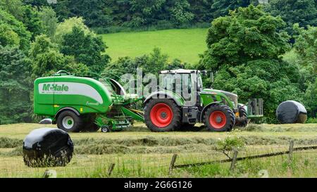 Production de foin ou d'ensilage (agriculteur travaillant dans un tracteur agricole sur une ramasseuse-presse à balles pour le travail dans les champs ruraux, ramassage de l'herbe sèche et balles rondes enveloppées) - Yorkshire England, Royaume-Uni. Banque D'Images