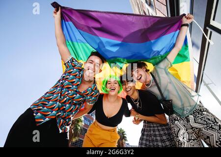 Quatre jeunes célèbrent leur identité queer lors d'un défilé de fierté gay. Groupe de jeunes LGBTQ+ souriant gaiement tout en élevant l'arc-en-ciel Banque D'Images