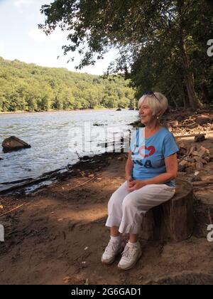 Belle femme blonde aux cheveux assis sur une souche le long du fleuve Delaware en Pennsylvanie tout en appréciant le paysage. Banque D'Images