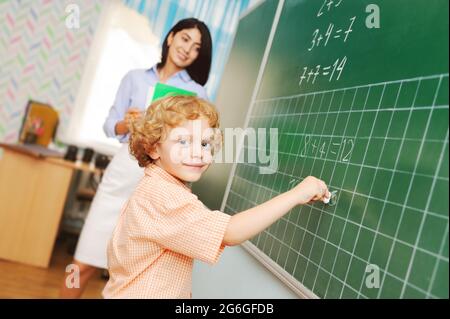 un petit garçon d'école mignon avec des cheveux bouclés écrit avec de la craie sur le tableau noir et décide un exemple mathématique avec une enseignante sur Banque D'Images