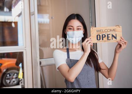 Le propriétaire d'un café asiatique portant un masque, une femme tenant l'enseigne pour ouvrir après avoir verrouillé la ville, prête à vendre et à travailler Banque D'Images