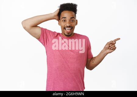 Portrait de beau afro-américain homme en t-shirt rose, toucher la tête et regarder surpris, le doigt pointant vers la droite à la vente logo, montrant la bannière, blanc Banque D'Images