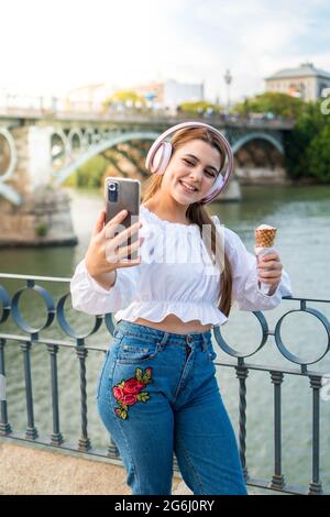 Jeune femme à l'écoute de musique, à manger une glace à l'extérieur en été et à prendre une photo d'elle-même. Femme caucasienne utilisant le téléphone portable et appréciant le Banque D'Images